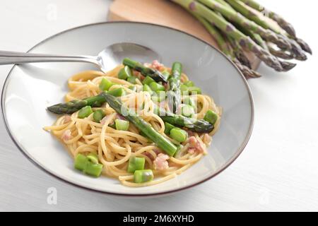 Spaghetti mit grünem Spargel, Speck und Ei als Variation der Carbonara-Sauce auf einem grauen Teller und auf einem weiß bemalten Holztisch, ausgewählter Fokus, schmal Stockfoto