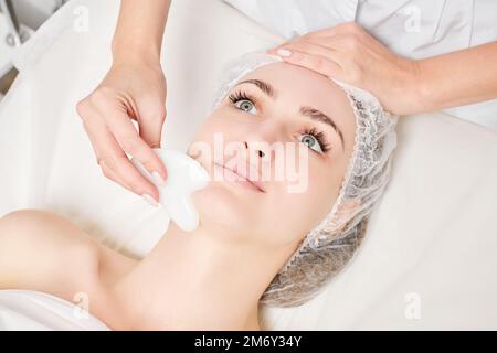 Kosmetologe macht Gesichtsmassage mit Gua Sha Stein der weiblichen Gesichtshaut für Lymphdrainage, Anti-Aging-Hautpflege kosmetische Prozedur im Beauty Spa Stockfoto