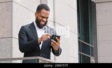 Männlicher, ethnisch erfolgreicher, lächelnder, bärtiger afroamerikanischer Unternehmer, Unternehmer, der mit dem Sieg des Mobiltelefons zufrieden ist, mit dem gute Nachrichten gewinnen Stockfoto