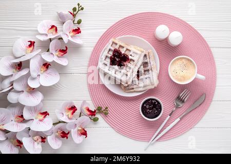Belgische Waffeln mit Latte und Blüten auf einem weißen Holztisch. Draufsicht. Muttertagskarte. Hintergrund für Speisen und Getränke. Kaffee, Nachtisch auf Brot Stockfoto