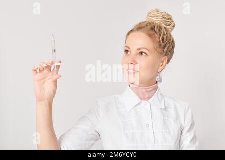 Eine Kosmetikerin in weißem Mantel hält die Spritze bereit, um eine Verjüngungsspritze für eine Kundin zu verabreichen, eine schöne lächelnde Frau auf weißem Hintergrund Stockfoto