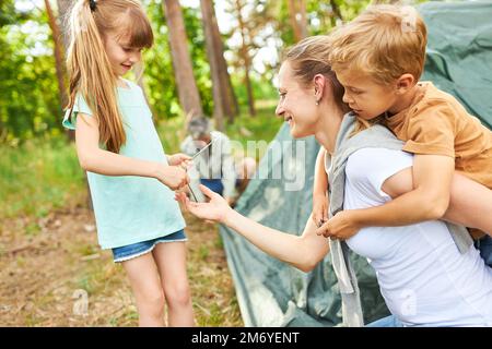 Ein Mädchen, das Mutter Huckepack-Sohn beim Campen im Wald im Urlaub einen Zeltpfahl gibt Stockfoto