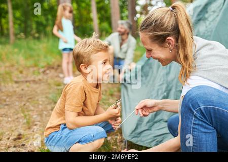 Ein Junge hält Haken und spricht mit der Mutter, während er während der Sommerferien ein Zelt im Wald aufbaut Stockfoto