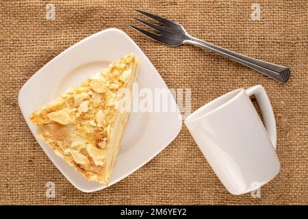 Ein Stück Cremetorte mit weißer Untertasse, Metallgabel und Tasse auf Jutetuch, Makro, Draufsicht. Stockfoto