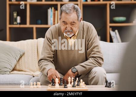 Senior man, denkt oder spielt Schach im Haus, Wohnzimmer oder Wohnung in japanischer Strategie, Schachmatt- oder Brettspielwettbewerb. Ruhestand Stockfoto