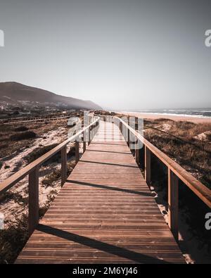 Eine lange Promenade auf einer grasbedeckten Sanddüne am Strand, die zu den Bergen und dem Ozean mit entsättigten Farben führt. Das Wegweiser-Konzept, Quiaios Beach Stockfoto