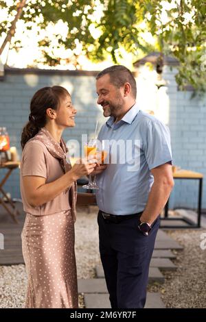 Ein glückliches Paar, das sich im Terrassengarten unterhalten und geflirtet hat und Cocktails serviert. Menschen, die bellini-Cocktails und Aperol Spritz zusammen in Outdoor s anstoßen Stockfoto