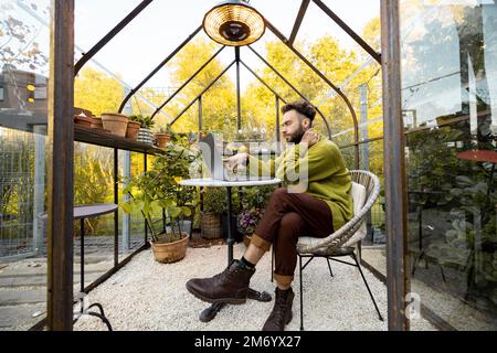 Mann arbeitet fernab auf Laptop im Glashaus im Hinterhof Stockfoto