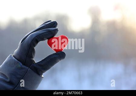 Rotes gestricktes Herz in weiblicher Hand in schwarzem Lederhandschuh auf Schnee- und Sonnenhintergrund. Konzept der romantischen Liebe, Valentinstag, Winterwetter Stockfoto