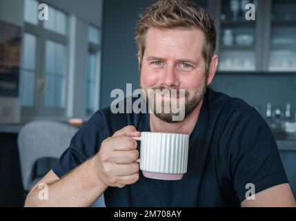 Nahaufnahme eines lächelnden jungen bärtigen Mannes, der Tee aus einer Tasse im Wohnzimmer trinkt Stockfoto