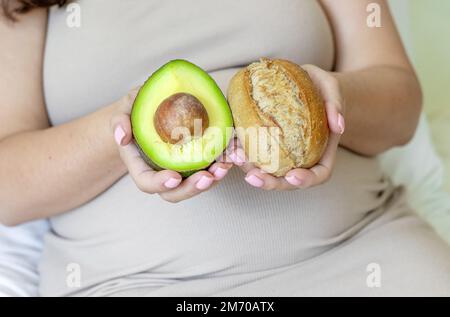 Schwangere Frau im Bett fortgeschrittener Bauch mit Avocado Brötchen Babykleidung in den Händen oder Creme Lotion auf den Arm auftragen, die in einem U-förmigen Kissen schläft Stockfoto