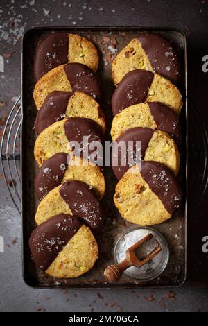 Ein Tablett mit Pistazien-Shortbread-Keksen, halb in dunkle Schokolade getaucht und mit Meersalz bestreut. Stockfoto