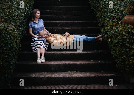 Ein romantisches Foto eines jungen Paares in einem wunderschönen Herbstpark, mit der Frau, die auf einer Treppe sitzt und dem Mann, der mit dem Kopf auf den Knien liegt Stockfoto
