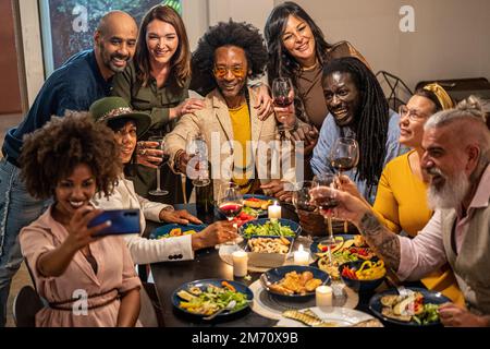 Verschiedene Menschen mit Alter und Ethnie essen ein veganes Abendessen. Multiethnische Gruppe von Freunden, die Spaß haben, während sie eine Mahlzeit in einem warmen und einladenden Hous teilen Stockfoto