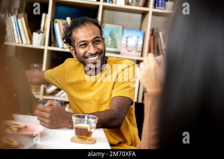 Lächelndes Porträt eines jungen Indianers in einer Bibliothek, junger Mann, der sich mit Freunden in der Cafeteria unterhält, Leute, die Tee trinken und Kekse essen Stockfoto