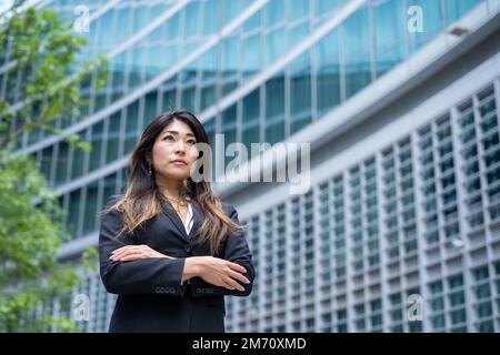 Selbstbewusste asiatische Geschäftsfrau mit gekreuzten Armen für ihre Leistungen, moderne Skyscreaper-Hinterwäldler, weibliche Geschäftsfrau japanischer Herkunft, Kopie Stockfoto