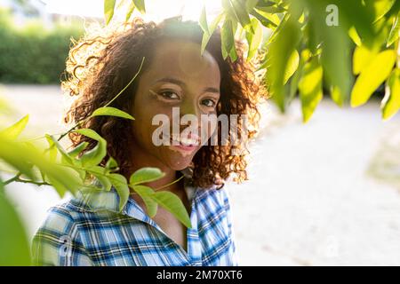 Porträt einer jungen birassischen Frau, die durch die Äste und Blätter eines Baumes geht, weibliche Person mit genetischer Vitiligo, die in die Kamera lächelt, außen Stockfoto
