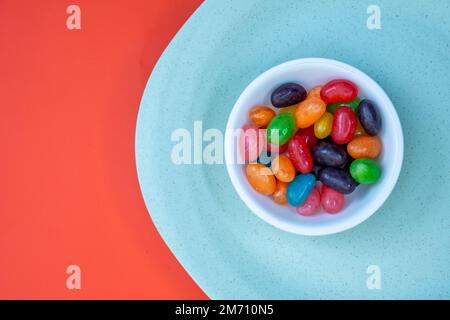 Weiße Schüssel mit Jelly Beans auf einer grünen Platte auf einem orangefarbenen Tisch, Designkonzept Stockfoto