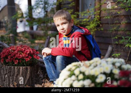 Ein süßer Kerl sitzt in einem Park, umgeben von weißen und roten Blumensträußen, und schaut mit einem leichten Grinsen in die Kamera. Modischer Teenager. Independen Stockfoto