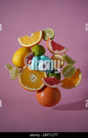 Ganze und geschnittene Früchte und die Spielzeugkamera schweben in der Luft auf einer rosa Nahaufnahme des Hintergrunds. Fliegende Orange, Limetten-Zitronen und Grapefruit. Magische Früchte. Frucht-sli Stockfoto