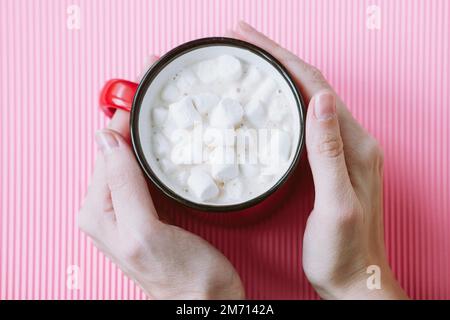Die Hände des Mädchens halten eine große Tasse Kakao oder heiße Schokolade mit Marshmallows auf pinkfarbenem Hintergrund, Draufsicht Stockfoto