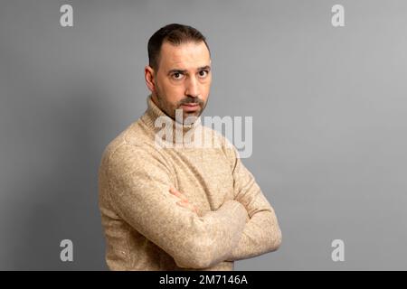 bärtiger hispanischer Mann in braunem Rollkragenpullover, der trotzig mit gekreuzten Armen steht und auf die Kamera schaut, isoliert über grau Stockfoto