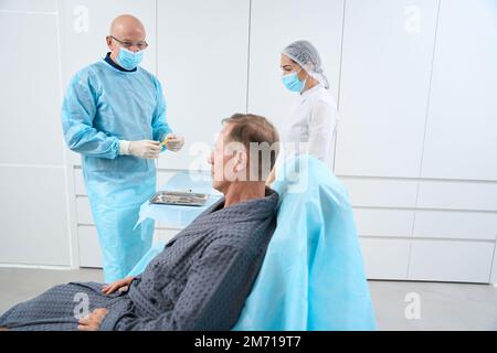 Glatzarzt in Operationsmaske und Handschuhen, die sich auf die Behandlung seines Patienten vorbereiten Stockfoto