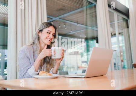 Junge Frau, die sich amüsiert, ihren Laptop benutzt, Kaffee aus einer Tasse trinkt. Angewohnheiten gegen Depressionen Stockfoto