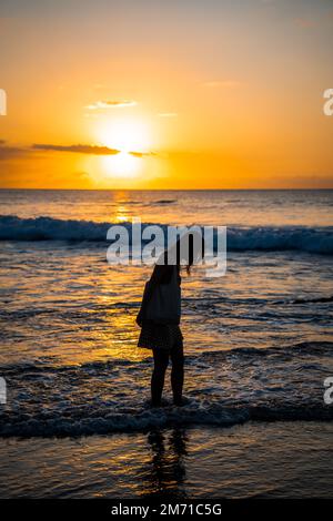 Ein vertikales Bild einer jungen Frau, die zur goldenen Stunde im Meer steht Stockfoto