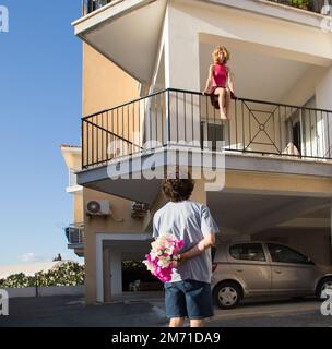 Der Typ will seinem geliebten Mädchen, das auf dem Balkon sitzt, einen Blumenstrauß schenken. Valentinstag-Überraschung. Konzept einer glücklichen romantischen Beziehung von Stockfoto