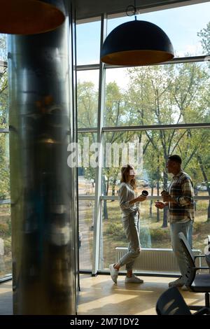 Mann und Frau halten Kaffee, während sie neben einem großen Fenster stehen Stockfoto