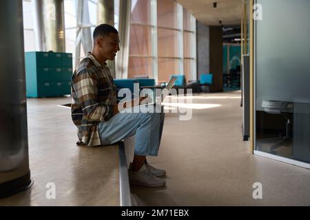 Lächelnder afroamerikanischer Mann, der auf einem Laptop tippt, während er auf einer Treppe sitzt Stockfoto