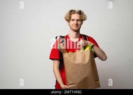 Lieferkonzept. Ein hübscher junger Liefermann in roter Uniform, der eine Packung Lebensmittel und Getränke aus dem Laden trägt, auf dem weißen Studio vermarktet Stockfoto