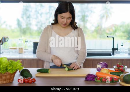 Schwangere Asiatin schneidet Gurke für frischen grünen Salat, weibliche bereitet leckeres Bio-Abendessen zu Hause zu, gesunde Ernährung für zukünftige Mutter Stockfoto