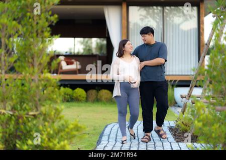 Schwangere Asiatin mit ihrem Mann, die zu Hause im Garten spazieren, um sich zu entspannen und frische Luft zu atmen. Stockfoto