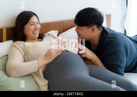 Porträt einer asiatischen, glücklichen schwangeren Frau und ihres Mannes, der Babyschuhe auf dem Bett im Schlafzimmer zu Hause hält Stockfoto