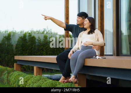 Schwangere Asiatin mit ihrem Mann, die sich zu Hause auf der Terrasse unterhalten, um zu entspannen und frische Luft zu atmen. Stockfoto