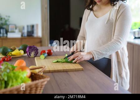 Schwangere Asiatin schneidet Gurke für frischen grünen Salat, weibliche bereitet leckeres Bio-Abendessen zu Hause zu, gesunde Ernährung für zukünftige Mutter Stockfoto