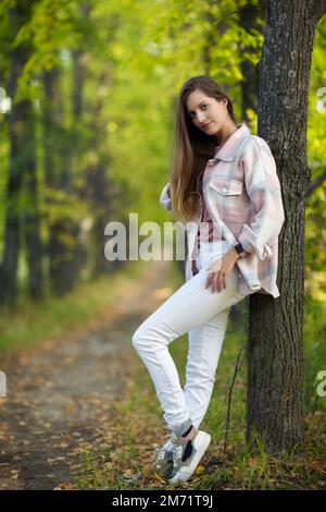 Blonde Frau in blauem Kleid, die sich in einer Herbstlindenallee an einen Baumstamm lehnt. Pfad unter gelben Bäumen mit herbstlichen Blättern. Sonniger Herbst Stockfoto