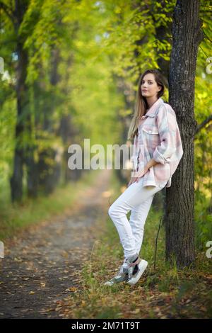 Blonde Frau in blauem Kleid, die sich in einer Herbstlindenallee an einen Baumstamm lehnt. Pfad unter gelben Bäumen mit herbstlichen Blättern. Sonniger Herbst Stockfoto