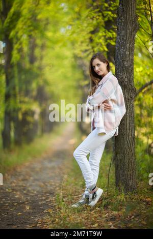 Blonde Frau in blauem Kleid, die sich in einer Herbstlindenallee an einen Baumstamm lehnt. Pfad unter gelben Bäumen mit herbstlichen Blättern. Sonniger Herbst Stockfoto