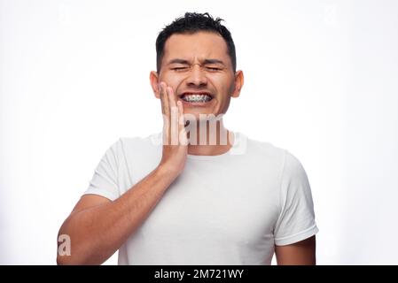 Ein hispanischer Mann in weißem Hemd und hält Grimaces in Schmerzen im Gesicht und hält sein Gesicht an seinen kieferorthopädischen Halterungen, eine Person Stockfoto