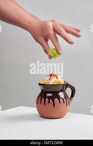 Ein Latino, ein lateinamerikanischer Mann presst eine grüne Limette über einen Esquite, Mais in einen Becher, ein typisches mexikanisches Gericht, in einen Tonbecher mit gelbem Mais, mexikanische Huasteca Stockfoto