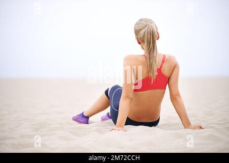 Wer braucht schon einen Fitnessraum, wenn man einen Strand hat? Rückansicht einer jungen Frau in Sportbekleidung, die am Strand sitzt. Stockfoto
