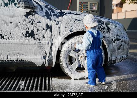 Rückansicht eines kleinen Jungen in blauem Overall und Kappe, der in der Autowaschanlage neben dem Seifenwagen steht und dabei beobachtet, wie weißer Schaum aus der Karosserie tropft. Stockfoto