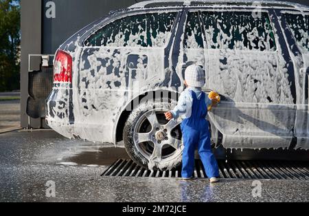 Rückansicht eines kleinen Jungen in Overall und Mütze, der mit Schwamm in den Händen neben der Seifenlauge der Karosserie steht. Kinderseife, die an der Autowäsche teilnimmt Stockfoto