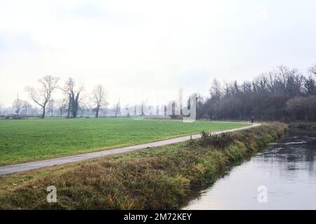 Asphaltierter Pfad mit einem Wald und einem Wasserstrahl am Rande inmitten von bebauten Feldern an einem bewölkten Tag in der italienischen Landschaft Stockfoto
