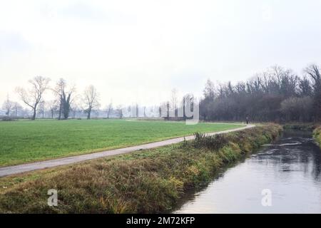 Asphaltierter Pfad mit einem Wald und einem Wasserstrahl am Rande inmitten von bebauten Feldern an einem bewölkten Tag in der italienischen Landschaft Stockfoto