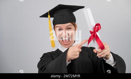 Glückliche Frau im Graduiertenkleid, die mit dem Finger auf ein Diplom zeigt. Stockfoto