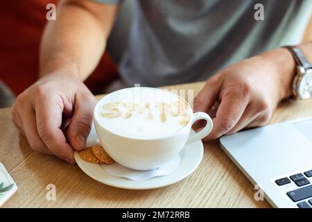 Gekürzte Hände halten die Kaffeetasse mit Sahne-Schaum SMM Zimt mit Keksen auf dem hölzernen Kaffeetisch. Social-Media-Marketing Stockfoto